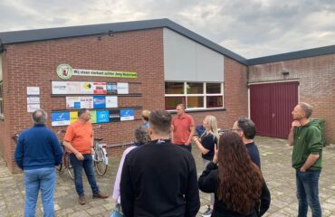 Leden van JAN staan voor het clubgebouw van Jong Nederland Budschop en krijgen uitleg van vrijwilligers