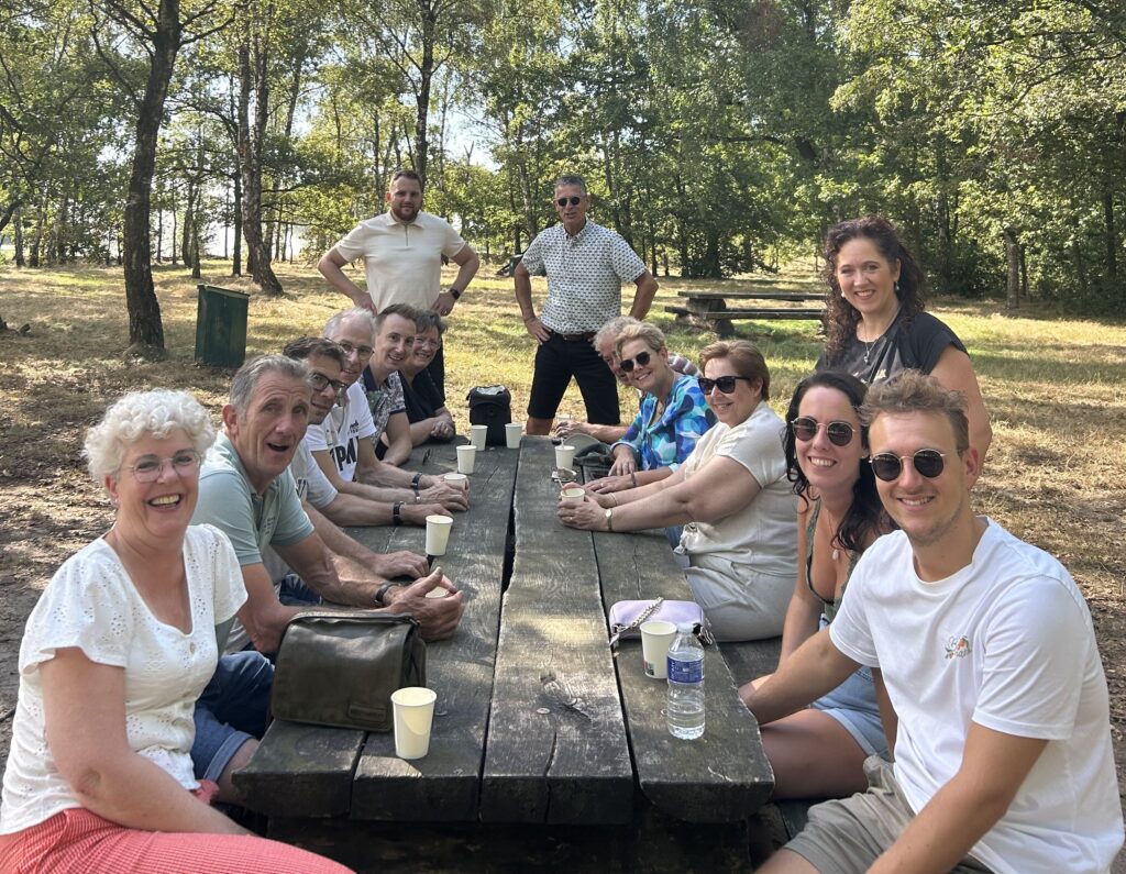 Leden van JAN zitten en staan lachend rond een picknicktafel terwijl ze genieten van een heerlijk stuk vlaai en een kopje koffie.