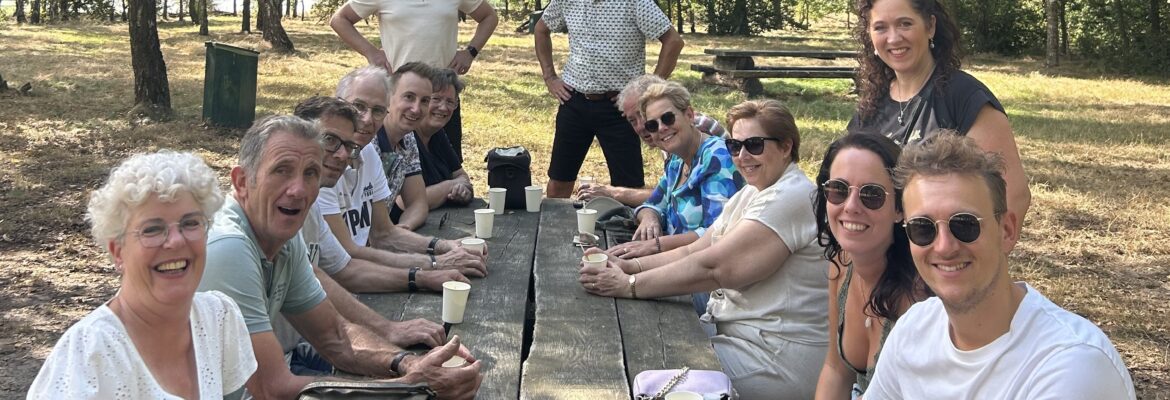 Leden van JAN zitten en staan lachend rond een picknicktafel terwijl ze genieten van een heerlijk stuk vlaai en een kopje koffie.