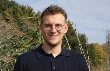 Lijsttrekker Kevin Bax staat buiten in de natuur en kijkt lachend naar de camera.