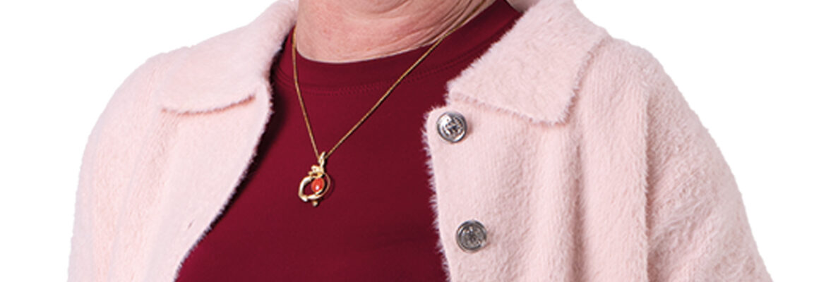 Carin Sieben staat met een hand in haar broekzak en kijkt je lachend aan. Ze draagt een bordeauxrood shirt met een roze vestje