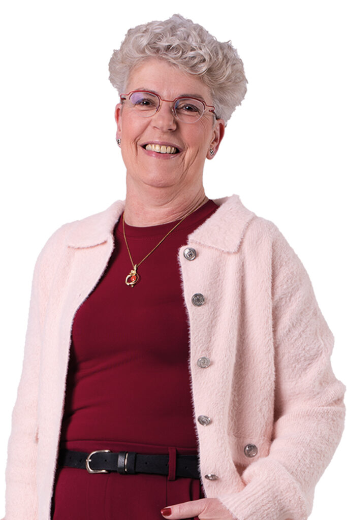 Carin Sieben staat met een hand in haar broekzak en kijkt je lachend aan. Ze draagt een bordeauxrood shirt met een roze vestje